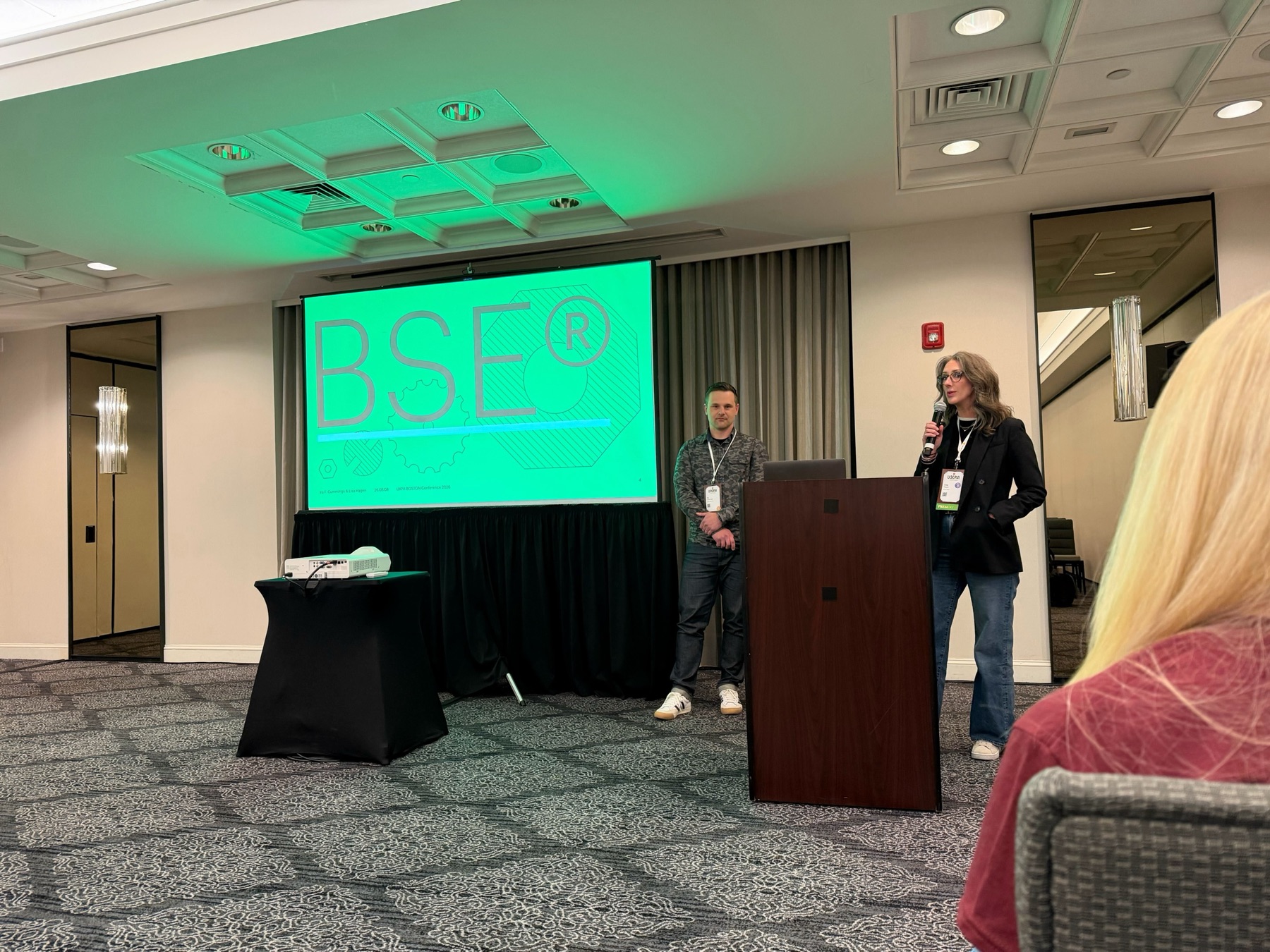 Lisa speaking at a podium beside a projected slide while Ira stands nearby at UXPA Boston.